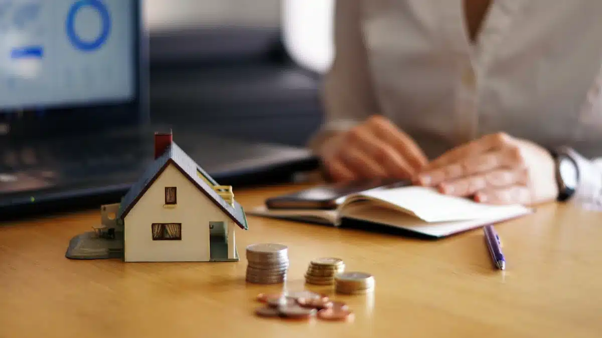 Close-up of a miniature house and coins on a desk, with a person reviewing financial documents in the background.